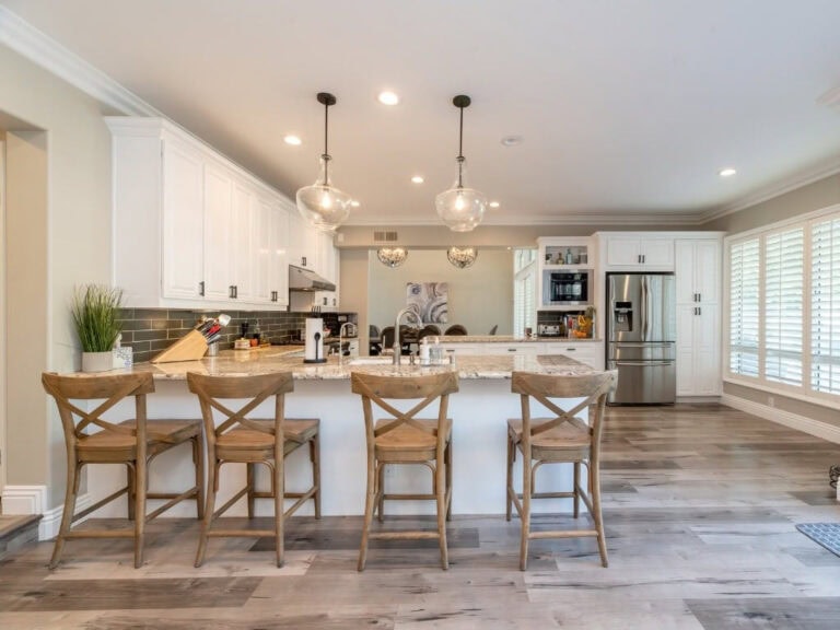 Modern kitchen with white cabinetry, stainless steel appliances, and a spacious island with four wooden bar stools, brightened by natural light from large windows.