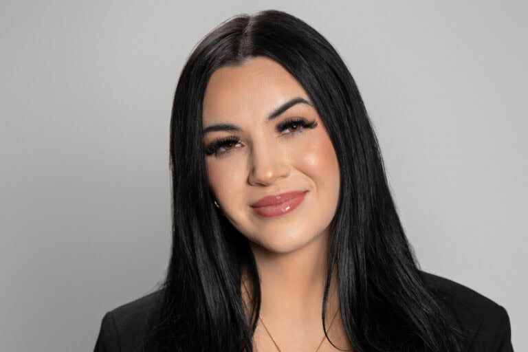 A professional Real Estate Agent, Jadyn Harbin, with long black hair and a confident smile, dressed in a black blazer, posing against a neutral gray background, representing professionalism and mastery in her field.