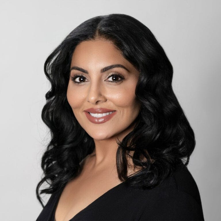 A professional Real Estate Agent, Komal Kaur Dhillon-Duran, with dark, wavy hair and a warm smile, dressed in a black top, posing confidently for a professional portrait.