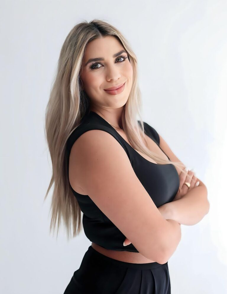 Confident young woman with long blonde hair smiling, wearing a black sleeveless top and black pants, standing with arms crossed against a white background.