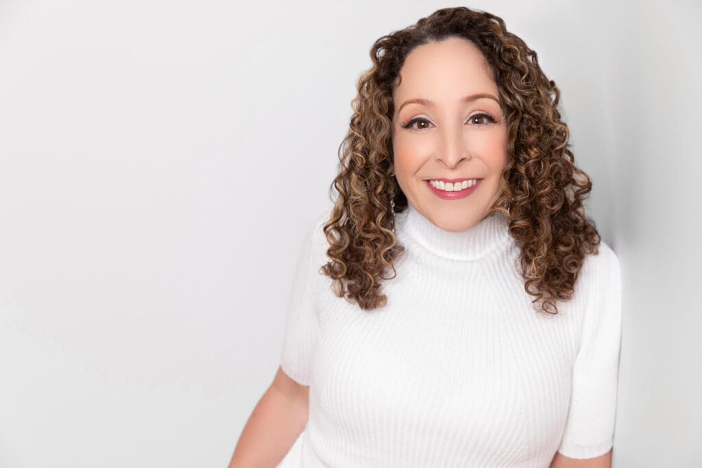 Smiling woman with curly hair wearing a white turtleneck in a professional portrait.
