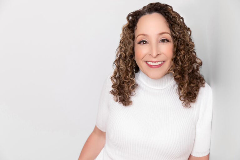 Smiling woman with curly hair wearing a white turtleneck in a professional portrait.