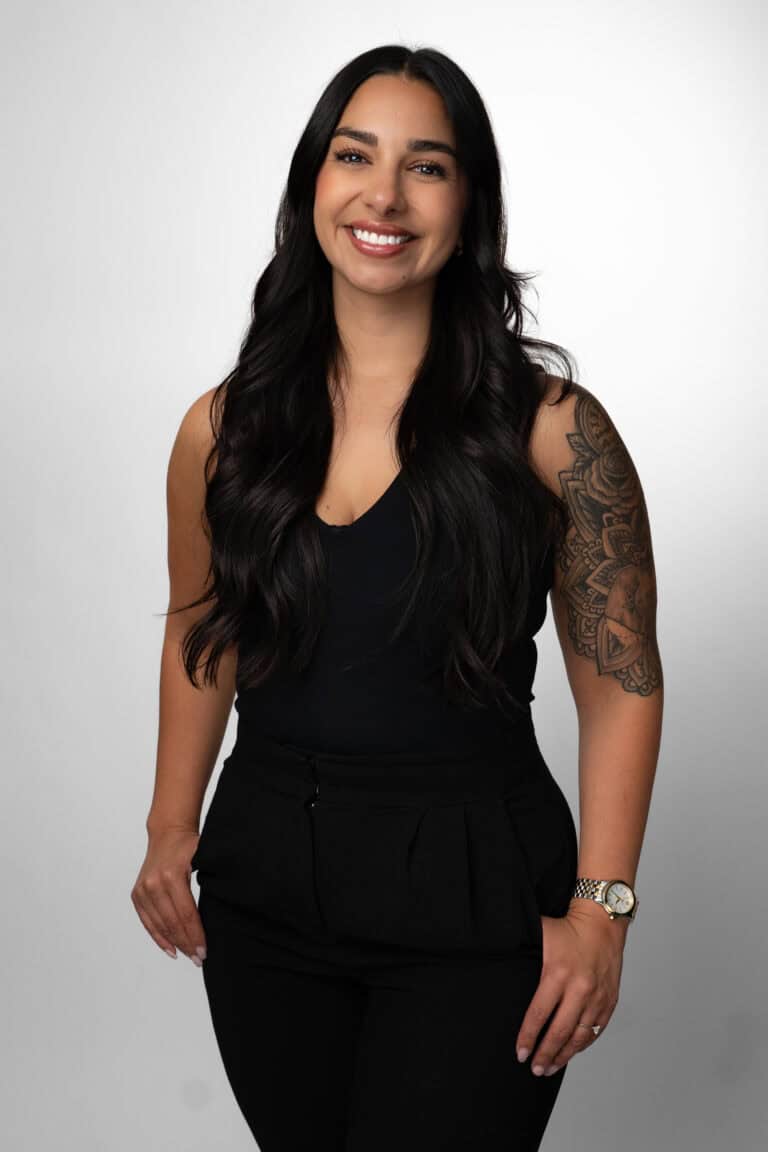 Portrait of a smiling woman with long dark hair, wearing a black sleeveless top and black pants, standing against a white background.